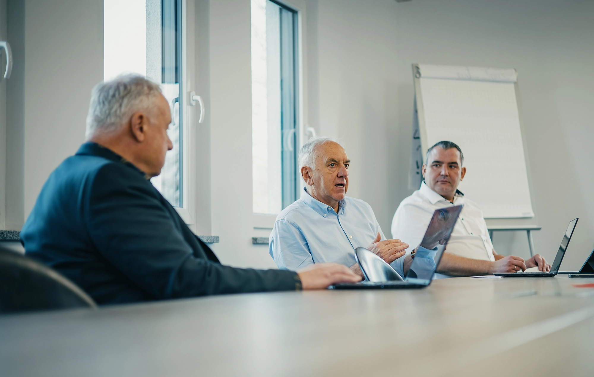 Drei Männer in einem Meetingraum diskutieren am Tisch mit Laptops, Fenster im Hintergrund, Flipchart an der Wand.