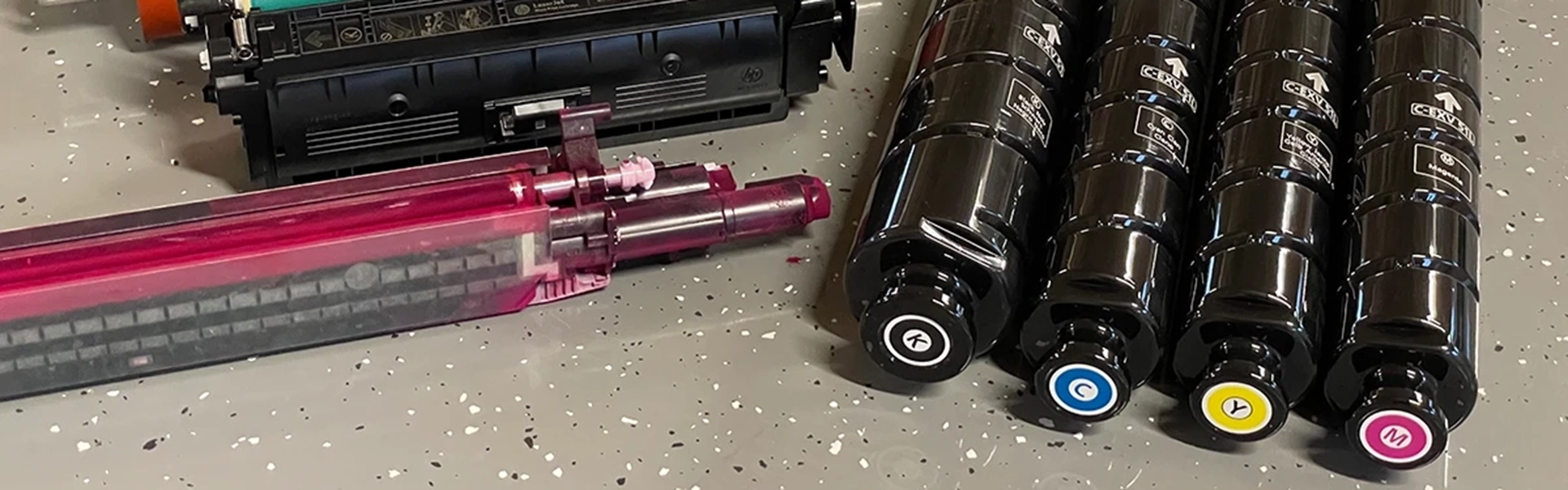 Vier Farblasertonerkartuschen in Schwarz, Cyan, Gelb und Magenta auf einem gesprenkelten Boden liegend.