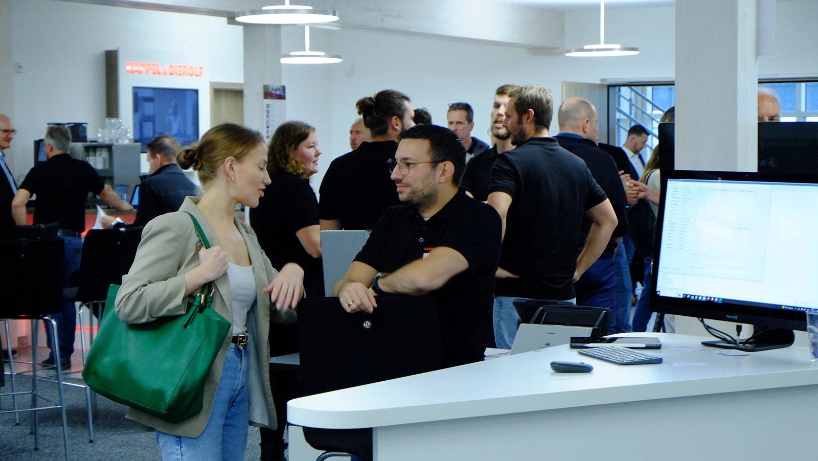 Zwei Personen unterhalten sich in einem modernen Büroraum mit vielen weiteren Gästen. Im Hintergrund sind Computer und ein Bildschirm sichtbar.