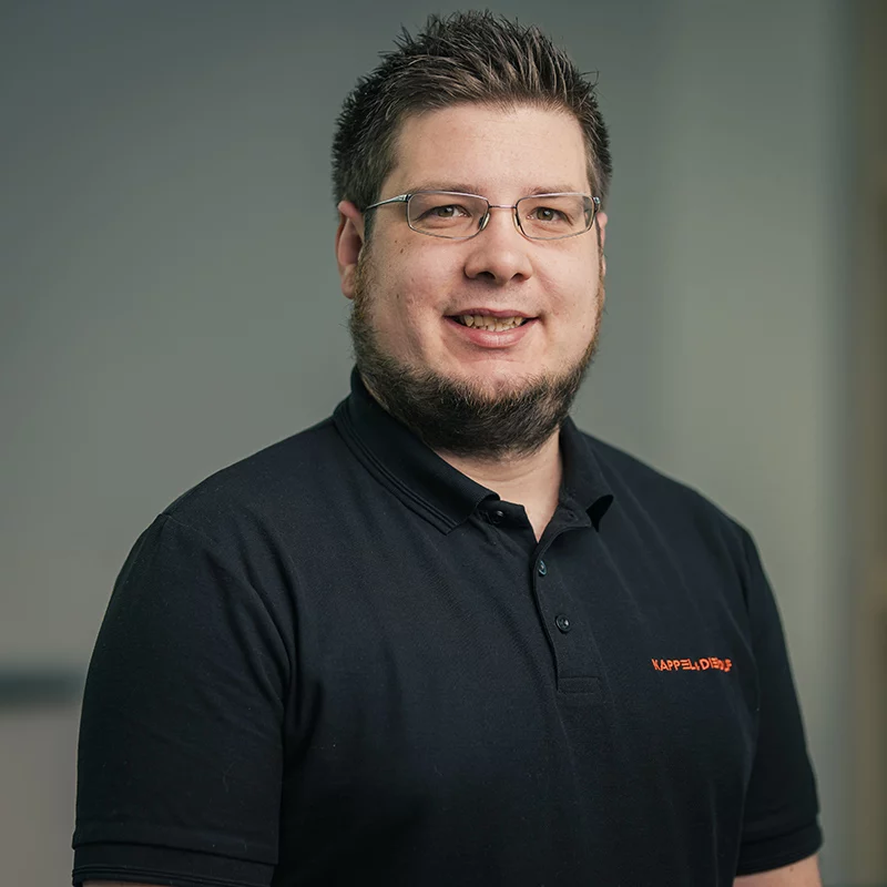 Mann mit Brille und Bart, trägt schwarzes Poloshirt, lächelt vor neutralem Hintergrund. Büroatmosphäre, freundliche Stimmung.