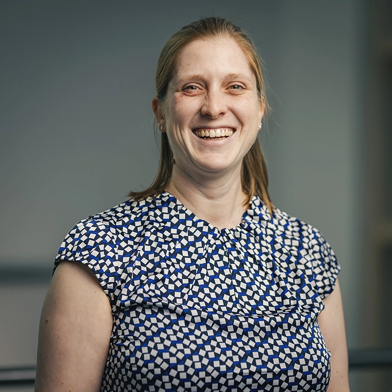 Frau mit blonder, schulterlanger Haar steht lächelnd vor neutralem Hintergrund. Sie trägt ein geometrisches Oberteil in Blau und Weiß.