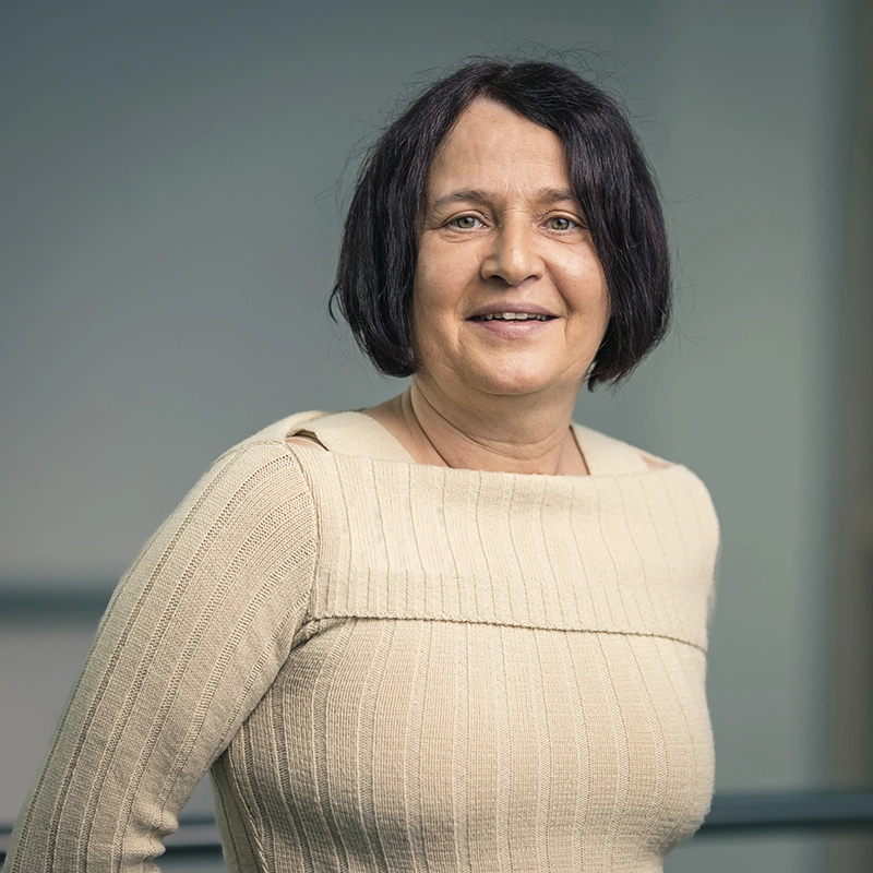 Porträt einer lächelnden Frau mit schulterlangen, dunklen Haaren, in einem beige gestreiften Oberteil, vor neutralem Hintergrund.