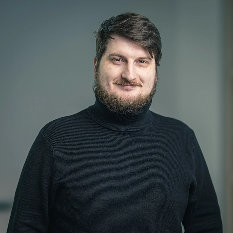 Mann mit blondem Bart und kurzem Haar, trägt einen schwarzen Rollkragenpullover, steht vor grauem Hintergrund und lächelt.