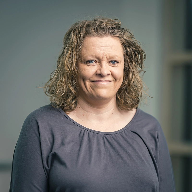 Porträt einer lächelnden Frau mit lockigem Haar in einem grauen Oberteil, vor einem neutralen Hintergrund.